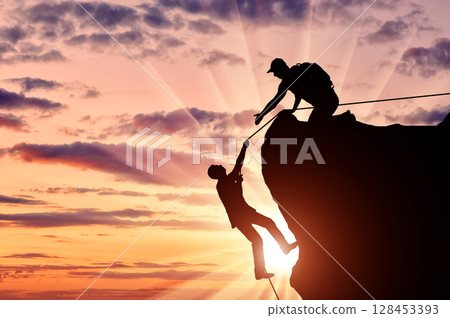 Silhouette of a climber who helps to climb the top of a man, throws him a rope and holds out his hand. 128453393