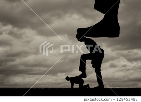 Silhouette of a large leg presses on a man who also presses his foot on another man lying on the ground 128453485