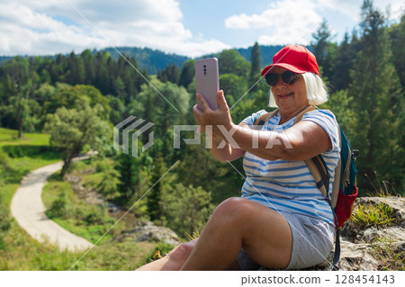 Smiling senior woman taking a selfie while hiking in the mountains 128454143