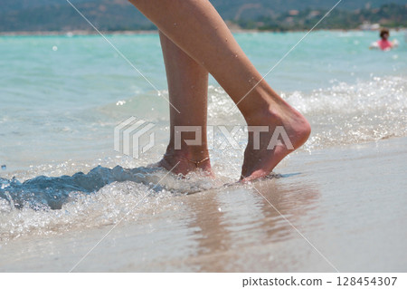 Close-up of female legs walking barefoot along the beach with sea waves. Wellness, travel and lifestyle concept Close-up of female legs walking barefoot along the beach with sea waves. Wellness, travel and lifestyle concept 128454307
