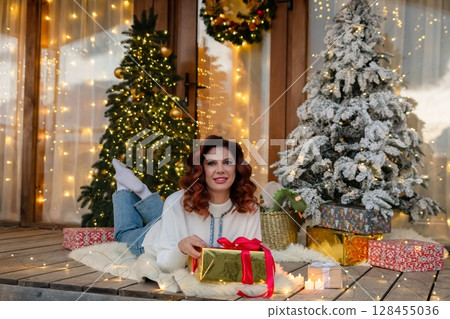 Woman Christmas Gift Decorated Porch - Woman lies on a porch with a gift, surrounded by Christmas trees and decorations. 128455036
