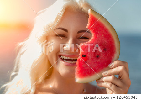 Woman Watermelon Summer Beach: Happy blonde enjoys juicy watermelon slice near ocean during sunny summer day. 128455045