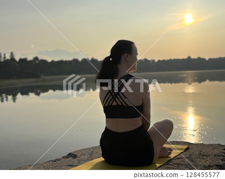 Woman sitting on fitness mat in yoga pose at sunrise by forest lake, back view. Outdoor workout, relaxation, mindfulness, healthy lifestyle, peaceful nature Woman sitting on fitness mat in yoga pose at sunrise by forest lake, back view. Outdoor workout, relaxation, mindfulness, healthy lifestyle, peaceful nature 128455577