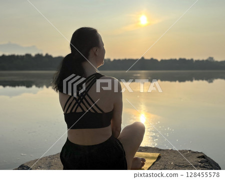 Woman sitting on fitness mat in yoga pose at sunrise by forest lake, back view. Outdoor workout, relaxation, mindfulness, healthy lifestyle, peaceful nature 128455578