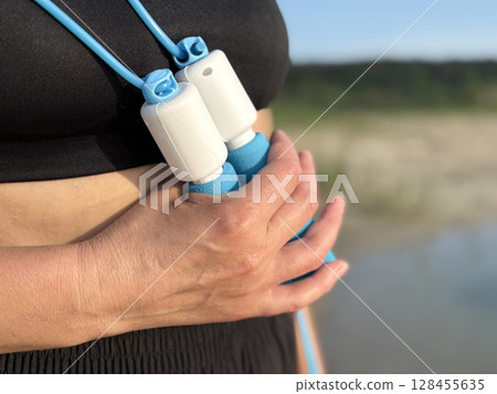 Athletic woman in black sportswear holding a blue skipping rope, standing in nature against the scenic backdrop of a forest and lake. Close up view emphasizing healthy lifestyle, outdoor fitness Athletic woman in black sportswear holding a blue skipping rope, standing in nature against the scenic backdrop of a forest and lake. Close up view emphasizing healthy lifestyle, outdoor fitness 128455635