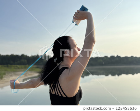 Athletic woman in black sportswear holding a blue skipping rope, standing in nature against the scenic backdrop of a forest and lake. Close up view emphasizing healthy lifestyle, outdoor fitness 128455636