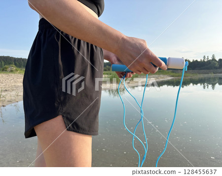 Athletic woman in black sportswear holding a blue skipping rope, standing in nature against the scenic backdrop of a forest and lake. Close up view emphasizing healthy lifestyle, outdoor fitness Athletic woman in black sportswear holding a blue skipping rope, standing in nature against the scenic backdrop of a forest and lake. Close up view emphasizing healthy lifestyle, outdoor fitness 128455637