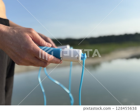 Athletic woman in black sportswear holding a blue skipping rope, standing in nature against the scenic backdrop of a forest and lake. Close up view emphasizing healthy lifestyle, outdoor fitness 128455638