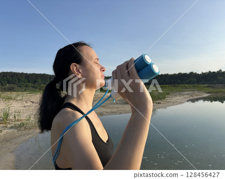 Athletic woman in black sportswear holding a blue skipping rope, standing in nature against the scenic backdrop of a forest and lake. Close up view emphasizing healthy lifestyle, outdoor fitness Athletic woman in black sportswear holding a blue skipping rope, standing in nature against the scenic backdrop of a forest and lake. Close up view emphasizing healthy lifestyle, outdoor fitness 128456427