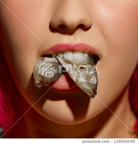 Close-up of woman with pink lipstick holding two fish in mouth against blue background Close-up of woman with pink lipstick holding two fish in mouth against blue background 128456549