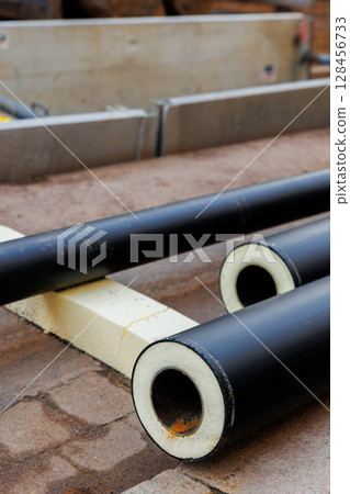 Heap of many new black insulated steel pipes at municipal construction site outdoors. Heating main district pipeline renewal or reconstruction. City development building industrial background 128456733