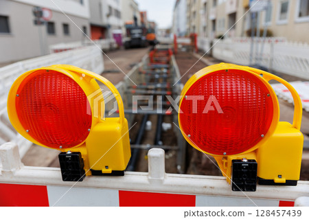 Urban street excavation with visible pipelines and safety barriers. Red warning lights and construction fencing mark the site in a residential area under infrastructure repair Urban street excavation with visible pipelines and safety barriers. Red warning lights and construction fencing mark the site in a residential area under infrastructure repair 128457439