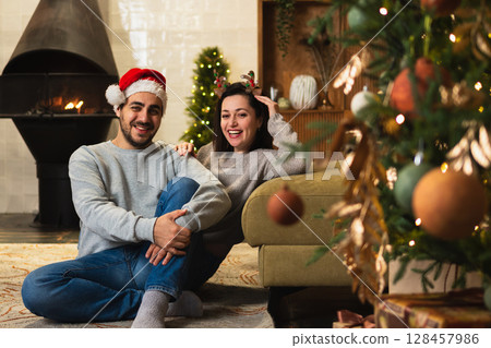 Merry Christmas. Happy young adult couple hugging bonding near Christmas tree and fireplace. Happy family man woman at home. People together celebrating Christmas enjoying winter holidays at home 128457986