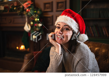 Merry Christmas. Young adult woman smiling near Christmas tree in classical dark interior. Happy girl in Santa hat in living room with fireplace Christmas tree gift boxes. Christmas eve at home Merry Christmas. Young adult woman smiling near Christmas tree in classical dark interior. Happy girl in Santa hat in living room with fireplace Christmas tree gift boxes. Christmas eve at home 128457999
