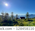 Mountain landscape with sunlit trees and distant cabin. 128458110