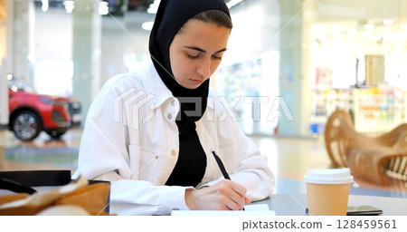 Focused muslim woman wearing hijab taking notes in notebook while sitting at table in shopping mall food court, enjoying a moment of productivity amidst the bustling atmosphere Focused muslim woman wearing hijab taking notes in notebook while sitting at table in shopping mall food court, enjoying a moment of productivity amidst the bustling atmosphere 128459561