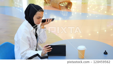 Professional businesswoman wearing traditional hijab sitting at table in shopping mall, recording voice messages on smartphone while working with pen and notebook in urban coffee environment Professional businesswoman wearing traditional hijab sitting at table in shopping mall, recording voice messages on smartphone while working with pen and notebook in urban coffee environment 128459562
