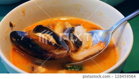 Savory tom yum soup brimming with succulent seafood, mussels, shrimp, squid, and mushrooms, carefully lifting from creamy white bowl with metal spoon Savory tom yum soup brimming with succulent seafood, mussels, shrimp, squid, and mushrooms, carefully lifting from creamy white bowl with metal spoon 128459564