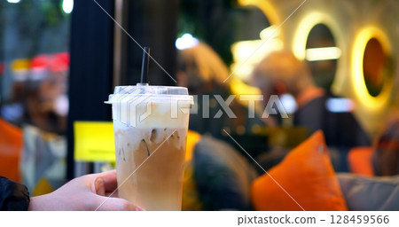 Barista presenting a delicious iced coffee with whipped cream and a straw, in a transparent plastic cup, with blurred customers and orange pillows in the background Barista presenting a delicious iced coffee with whipped cream and a straw, in a transparent plastic cup, with blurred customers and orange pillows in the background 128459566