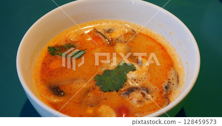 Traditional tom yum soup presenting vibrant flavors, featuring fresh mushrooms, tomatoes, and cilantro garnishing aromatic broth, nestled in white ceramic bowl against green background Traditional tom yum soup presenting vibrant flavors, featuring fresh mushrooms, tomatoes, and cilantro garnishing aromatic broth, nestled in white ceramic bowl against green background 128459573