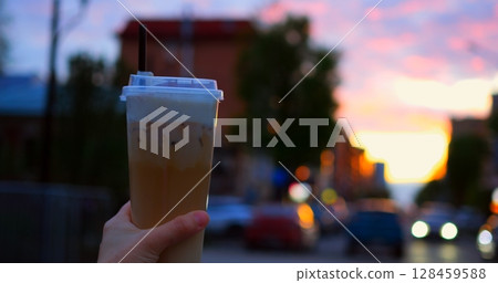 Hand gripping chilled coffee drink with whipped cream, straw visible against blurred urban sunset, background featuring traffic lights, city buildings 128459588