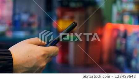 Close up of a customer's hand holding a smartphone, making a contactless payment at a store's checkout counter, with colorful blurred shelves in the background 128459648