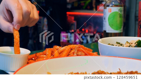 Restaurant patron dipping crispy sweet potato fry into vibrant green sauce, revealing casual dining experience with multiple dishes surrounding appetizer on restaurant table 128459651