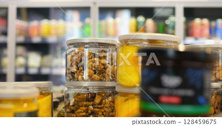 Transparent jars filled with walnuts and dried fruits are neatly arranged on a shelf in a supermarket, with other products visible in the background 128459675