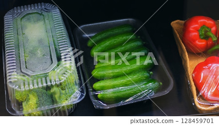 Fresh vegetables including cucumbers, bell peppers, tomatoes, and broccoli gradually appearing, wrapped in plastic film within organized supermarket produce display Fresh vegetables including cucumbers, bell peppers, tomatoes, and broccoli gradually appearing, wrapped in plastic film within organized supermarket produce display 128459701