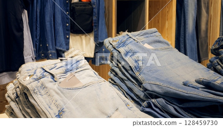 Multiple stacks of folded blue jeans sit on a wooden display table in a clothing store, surrounded by other apparel items, creating visually appealing display for shoppers. Concept fashion clothes Multiple stacks of folded blue jeans sit on a wooden display table in a clothing store, surrounded by other apparel items, creating visually appealing display for shoppers. Concept fashion clothes 128459730