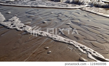 Ocean waves gently caress the sandy shore, creating mesmerizing patterns and textures as the water recedes, leaving behind a beautiful display of natural artistry 128460096