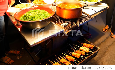 Southeast asian street food vendor preparing and grilling meat skewers, showcasing the cooking process alongside bowls of vegetables and soup illuminated by bright light bulbs 128460289