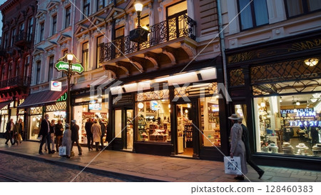 Elegant woman wearing a beige trench coat and hat walks past illuminated souvenir shops on a cobblestone street in lviv, ukraine, at dusk, while other pedestrians stroll along the sidewalk 128460383