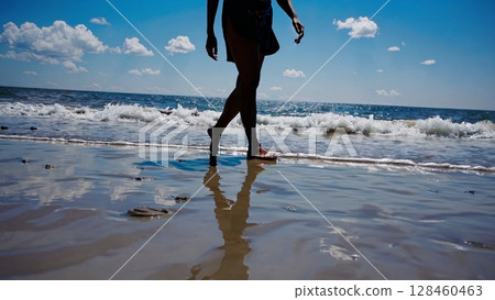 Tourist legs walking barefoot on glistening wet sand, leaving ephemeral footprints reflecting across shimmering water surface near ocean shoreline during sunny summer day 128460463