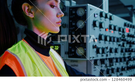 Female industrial worker monitoring control panel, managing machinery operations, ensuring smooth production process, and maintaining safety protocols in factory setting 128460499