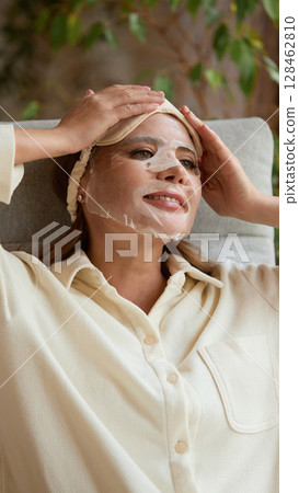 Night care ritual. Smiling mature woman with facial sheet mask relaxing on chair in beige shirt indoors 128462810