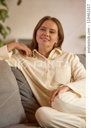 Joy of aging. Mature woman in cream loungewear sitting on couch smiling confidently in cozy living room 128462817