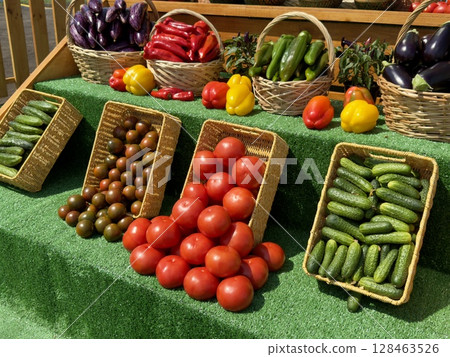 Red tomatoes, green cucumbers, black tomatoes, peppers, and eggplants are displayed in wicker baskets on a market stall, creating a colorful and inviting scene 128463526