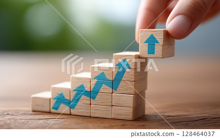 A hand adding wooden blocks to form an upward-trending graph, with blue arrows pointing up and the target at the top of the block representing progress towards business goals or growth. A hand adding wooden blocks to form an upward-trending graph, with blue arrows pointing up and the target at the top of the block representing progress towards business goals or growth. 128464037