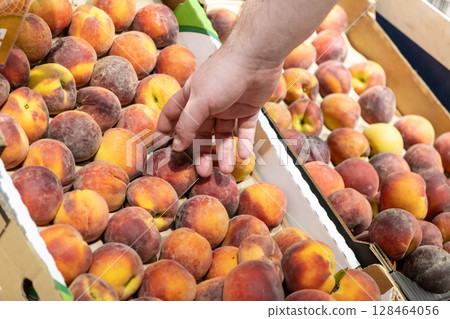 Person's hand picking a vibrant, ripe peach from a large, overflowing display of fresh fruit at an organic produce market, emphasizing healthy nutrition and consumer choice 128464056