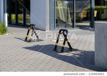 Two black and yellow foldable parking barriers with a red padlock secure reserved spots on a paved lot, ensuring private vehicle access control outside a modern office building 128464061