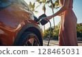 Woman charging electric vehicle at sunset in a tropical location with palm trees 128465031