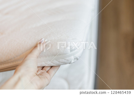 A hand gently pulls back a mattress protector to show the inner layer of the mattress. The setting suggests a focus on hygiene and maintenance in a well-lit bedroom environment 128465439