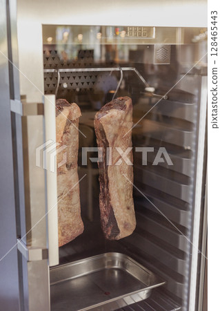 Two large cuts of beef are hung inside a modern refrigerator designed for dry aging. The meat appears well-marbled, and the environment is clean, emphasizing the butchering process 128465443