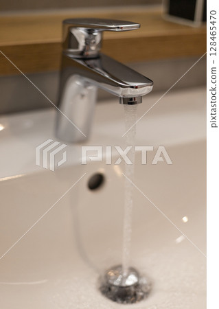 A sleek chrome faucet dispenses a steady stream of water into a pristine white sink, showcasing modern bathroom design with a minimalist aesthetic and clean lines 128465470