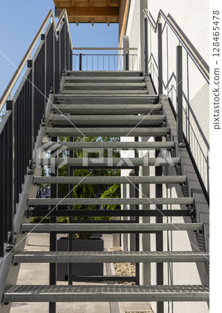A metal staircase with open risers ascends to a building's entrance. Sunlight illuminates the steps, highlighting their sturdy design. Metal construction, welding, steel stairs. A metal staircase with open risers ascends to a building's entrance. Sunlight illuminates the steps, highlighting their sturdy design. Metal construction, welding, steel stairs. 128465478