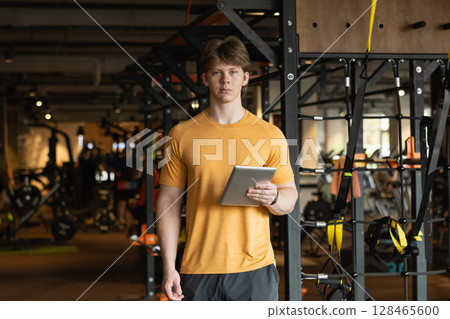 Personal trainer or bodybuilder standing wearing orange t-shirt holding tablet or clipboard in gym or fitness center. Coach making exercise plan in gym 128465600