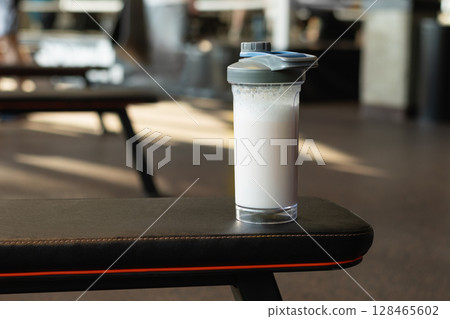 Protein shake cup resting on gym bench. Fitness enthusiasm and post-workout routine. Healthy lifestyle. Close up. 128465602