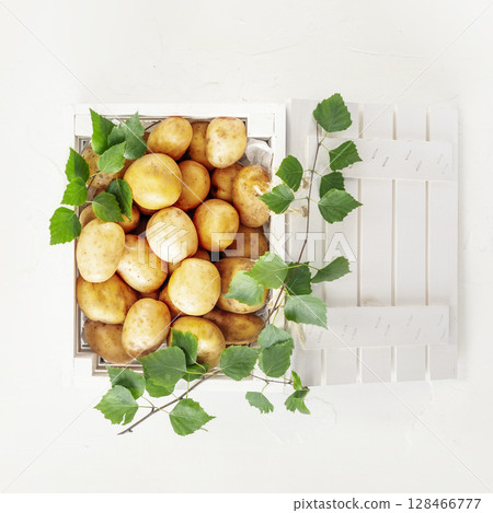 Raw potatoes. Natural, farm potatoes in a white wooden box with green leaves on white background. Selling seasonal vegetables at local market. Support local farmers and harvesting concept. Flat lay 128466777