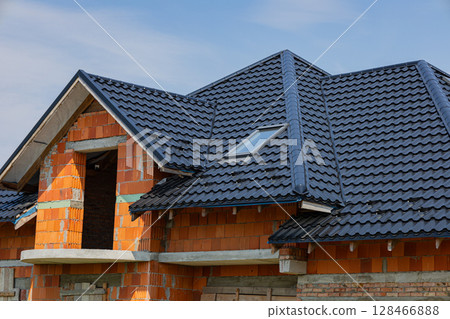 A metallic tiled roof is being installed on a residential building with brick walls, showcasing modern construction techniques A metallic tiled roof is being installed on a residential building with brick walls, showcasing modern construction techniques 128466888